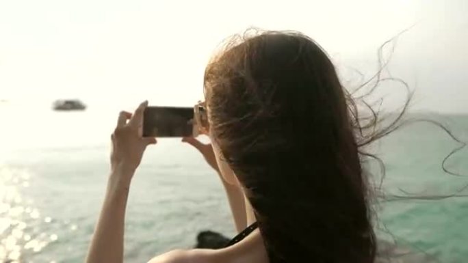 海滩岛上的女游客在乘船和天际线度假时用智能手机拍摄日落照片