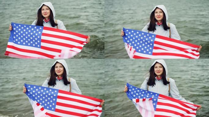 快乐的美国女孩的慢动作肖像举着美国国旗，并将其移动，站在海边，可见海浪。国籍和国家概念。