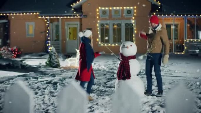 幸福的年轻夫妇在下雪的时候在他们田园诗般的房子的后院堆雪人。他们用围巾包裹雪人。家人在平安夜共度时光