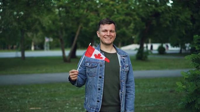 英俊的男性旅行者的慢动作肖像开朗的家伙挥舞着加拿大国旗，看着相机，微笑着站在美丽的城市公园里，树木和