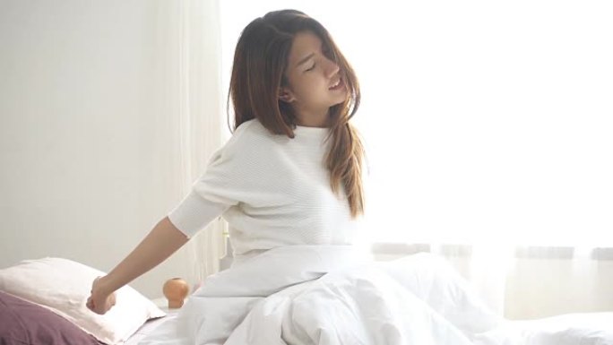 慢动作-快乐美丽的年轻亚洲女子早上醒来，坐在床上伸展舒适的卧室窗户。醒来后有趣的亚洲女人。亚洲女人在