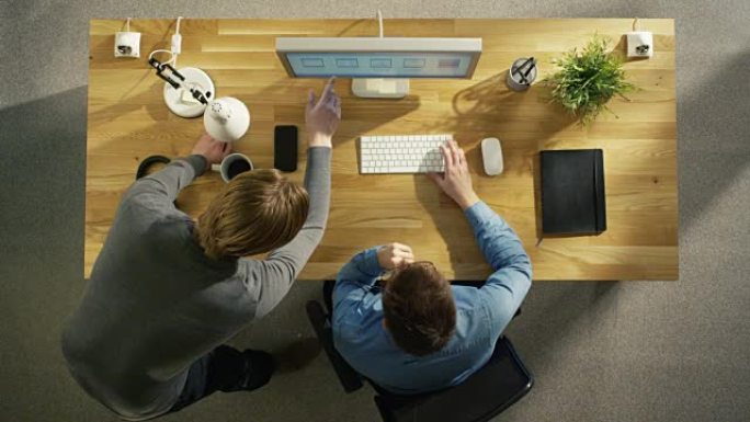 两位平面设计师讨论台式计算机工作的俯视图。桌子上还有: 笔记本，智能手机，耳机。