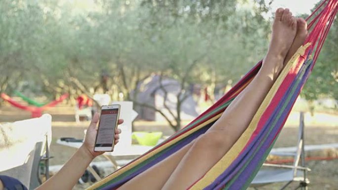 女人在吊床上放松，在夏季露营地使用智能手机