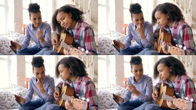 混合种族年轻女子平板电脑坐在床上教她十几岁的妹妹在家弹吉他
