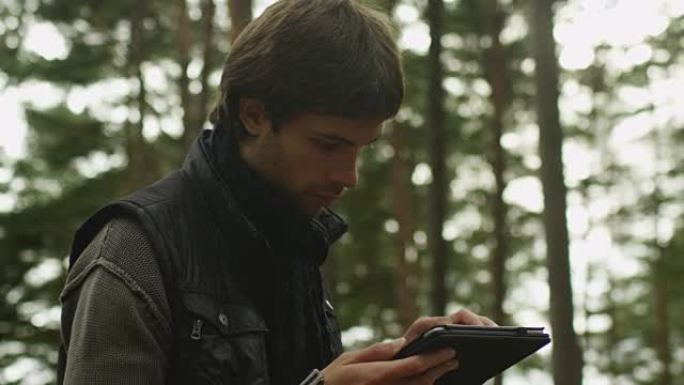 穿着深色秋衣和围巾的长发男子站在森林里，使用平板电脑。