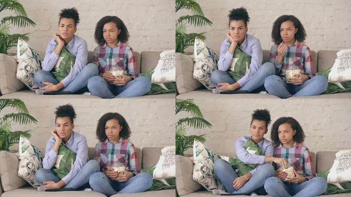 两个混血卷曲女孩朋友坐在沙发上看紧张的电视节目，在家吃爆米花