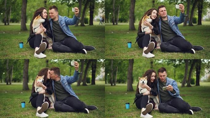 英俊的年轻人正在与抱着血统狗的女友自拍，盖伊在亲吻女孩，然后在屏幕上观看照片，人们在笑。