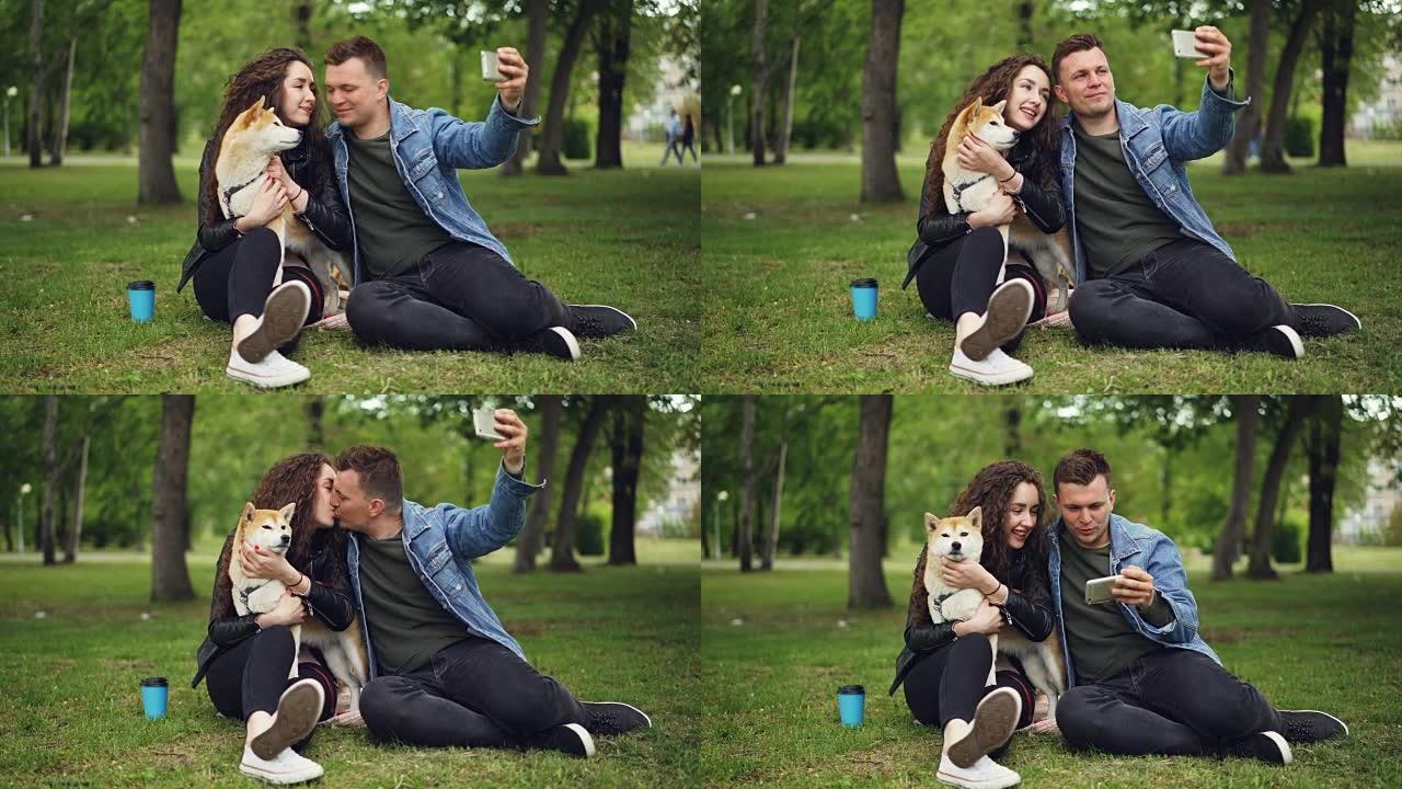 英俊的年轻人正在与抱着血统狗的女友自拍，盖伊在亲吻女孩，然后在屏幕上观看照片，人们在笑。