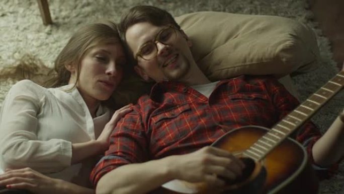 年轻快乐的微笑夫妇躺在地板上，晚上在家弹吉他。休闲生活方式。