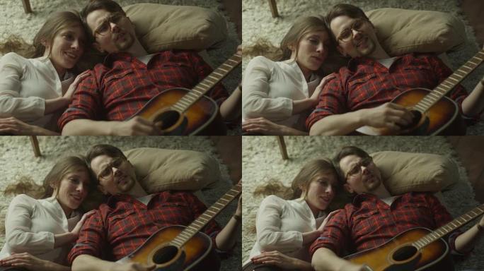 年轻快乐的微笑夫妇躺在地板上，晚上在家弹吉他。休闲生活方式。