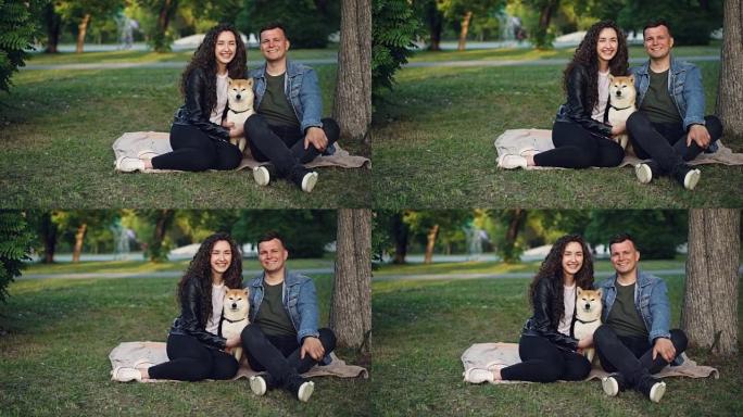 可爱的情侣和血统狗的慢动作肖像坐在公园草地上的毯子上，看着相机，微笑着。自然和家庭概念。