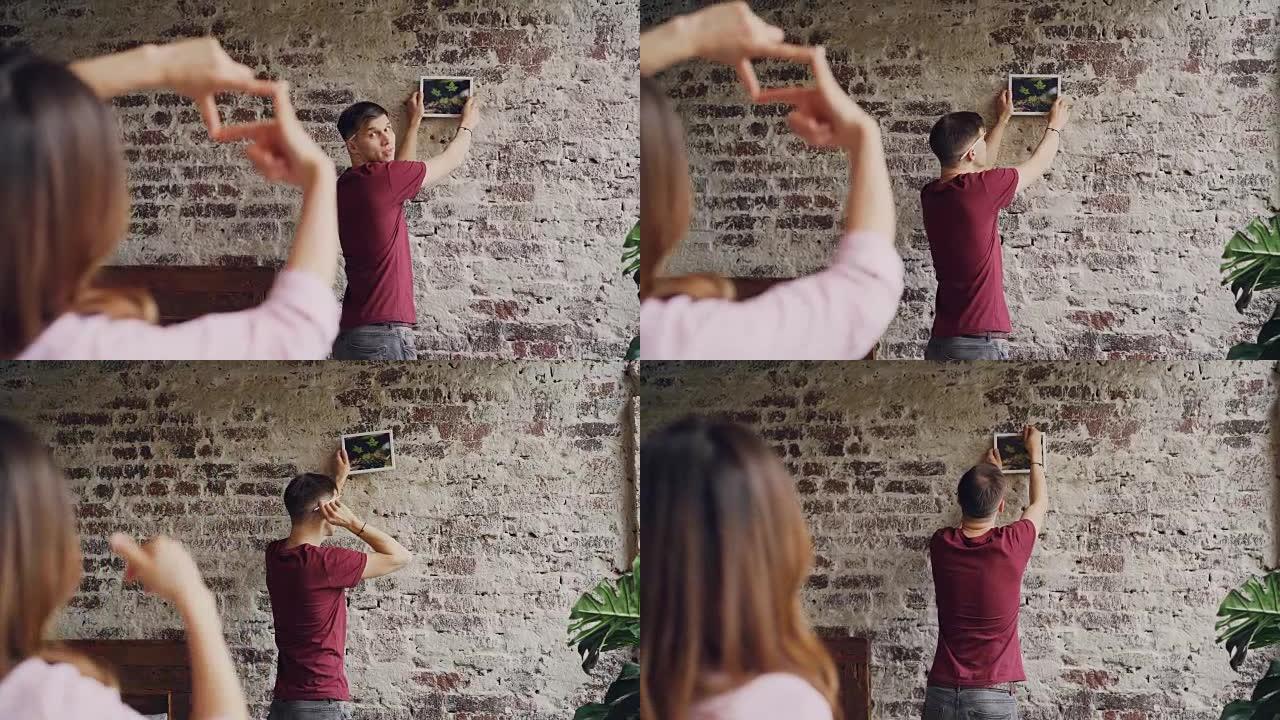 年轻的丈夫正在选择在砖墙上悬挂照片的位置，而妻子则用手指做框架，并示意以拇指表示他的决定。