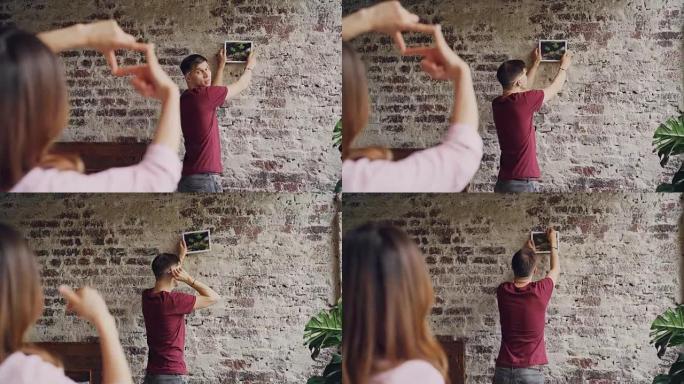 年轻的丈夫正在选择在砖墙上悬挂照片的位置，而妻子则用手指做框架，并示意以拇指表示他的决定。