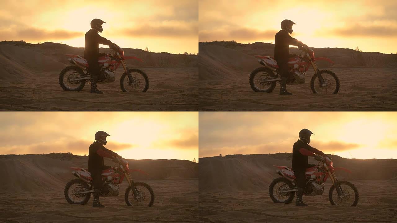 专业FMX摩托车骑手在他的摩托车上休息，俯瞰坚硬的沙地越野地形。太阳设置。