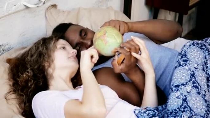快乐的多种族夫妇躺在床上扭动地球。男人和女人指向旅行的目的地，笑