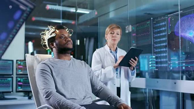 在实验室中，戴着脑电波扫描耳机的人坐在椅子上，而平板电脑的科学家则监督过程。在现代大脑研究/神经研究