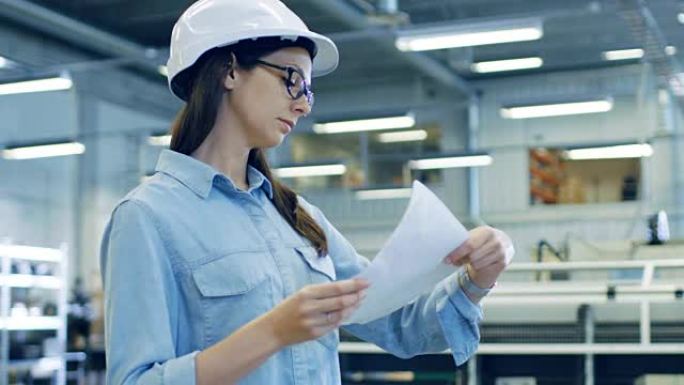 美丽的黑发工业工程师戴着安全帽，在穿过工厂时研究蓝图和文件。