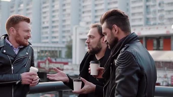 三个留着胡须的英俊年轻人在城市的桥梁栏杆附近进行交谈和喝咖啡。慢mo