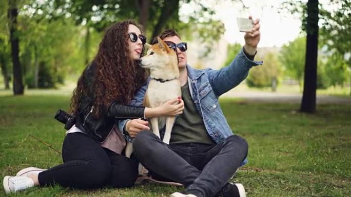 英俊的年轻人正在和他漂亮的妻子和可爱的狗自拍，他们都戴着墨镜。盖伊拿着智能手机拍照和摆姿势。