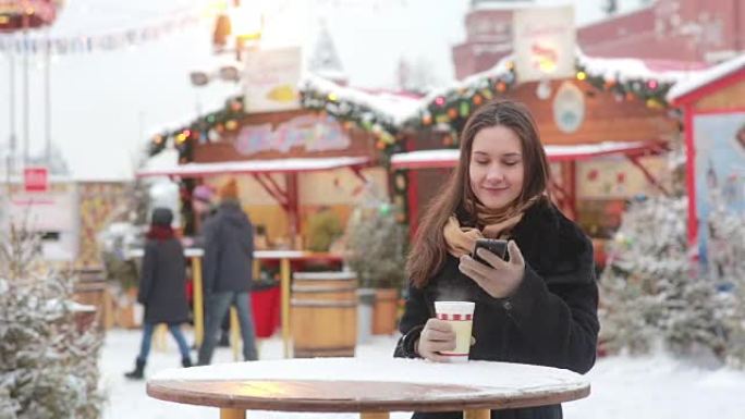 美丽的年轻女子在圣诞节博览会上使用智能手机并喝热茶