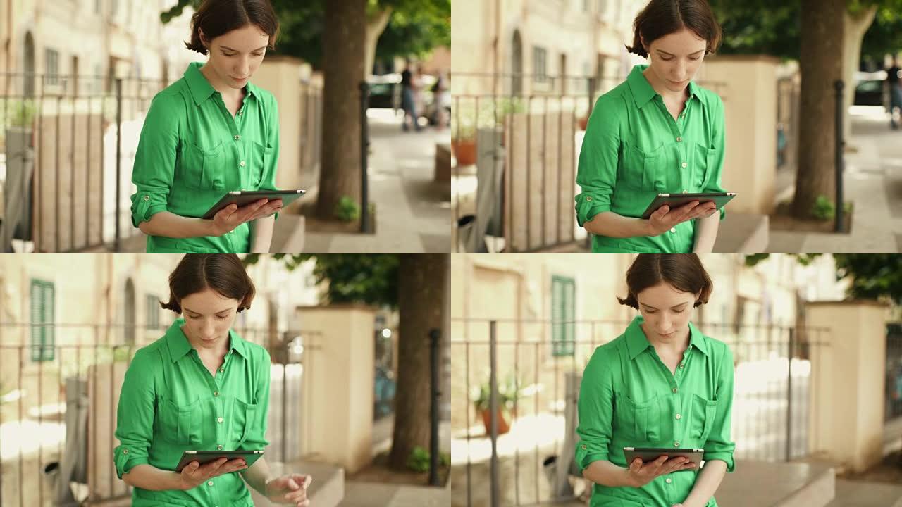 迷人的年轻女子穿着轻便的夏装，在欧洲小镇的街道上使用平板电脑。