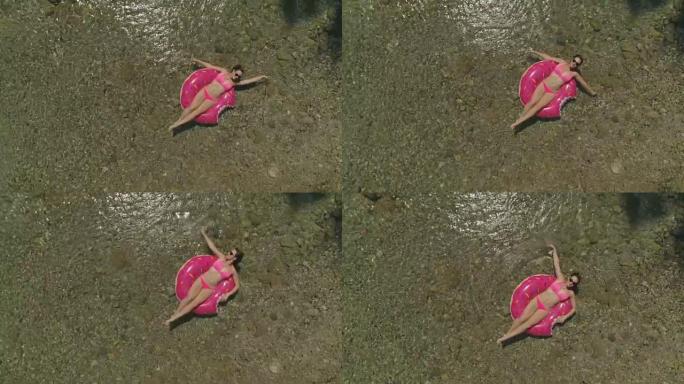 空中年轻女子在甜甜圈漂浮物上河边，双臂扫过水面