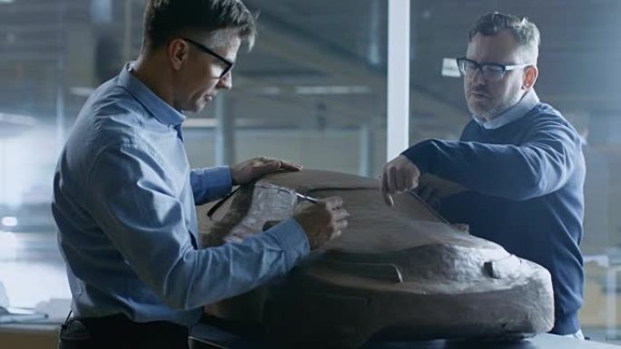 两位男性汽车设计师致力于新一代电动汽车未来设计的粘土模型。一个拿着平板电脑，另一个用耙子/电线用粘土