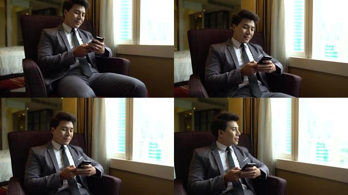 年轻的商人坐在酒店房间的椅子上，在他的智能手机移动技术连接运营商旅行设备小工具专业通信上输入一条消息