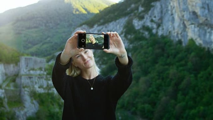 美丽的金发女孩用智能手机自拍的肖像，拿着屏幕对着相机。在背景风景秀丽的青山山景。