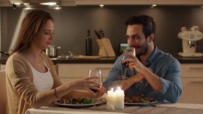 美丽的夫妇在厨房吃烛光晚餐。他们吃饭、喝酒和说话。为了庆祝他们碰杯。