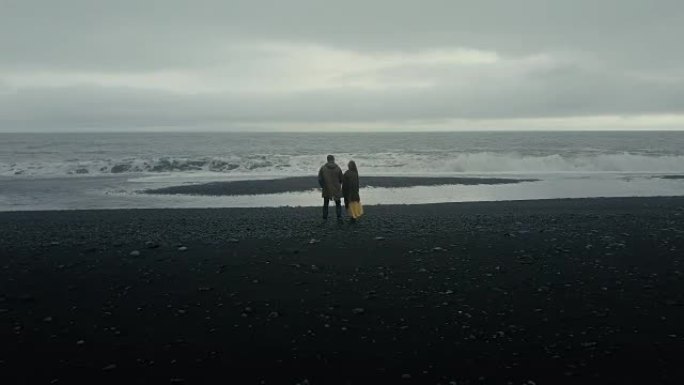 年轻夫妇站在黑火山海滩上，靠近巨魔脚趾岩石，在冰岛享受海浪的鸟瞰图