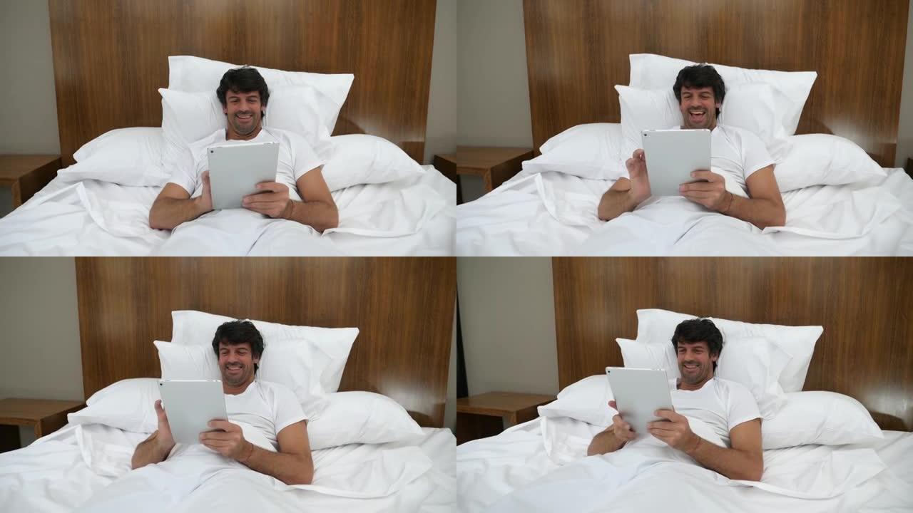 成年男子躺在家里的床上，用数字平板电脑聊天，看起来很开心，笑得很开心