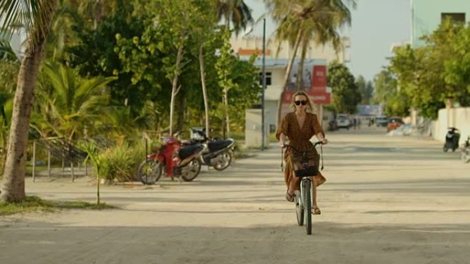 穿着连衣裙的美女骑着自行车在路上穿过正宗的小镇。南亚自然，风景秀丽的森林在路边生长。