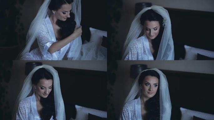年轻迷人的新娘穿着优雅的白色内衣，peignoir坐在床上。卧室里戴着面纱的漂亮女人
