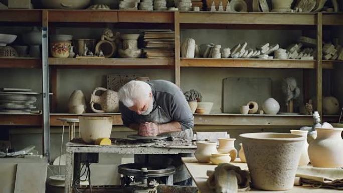 勤奋的银发男子正在陶工的轮子上使用粘土，塑造一片壤土。美丽的陶瓷器皿，手工制作的盆和架子上的花瓶是可