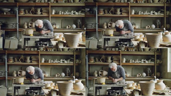 勤奋的银发男子正在陶工的轮子上使用粘土，塑造一片壤土。美丽的陶瓷器皿，手工制作的盆和架子上的花瓶是可