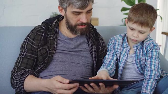 慈爱的父亲和他可爱的小孩正在使用桌子触摸屏，坐在家里的沙发上聊天。现代科技和幸福家庭理念。