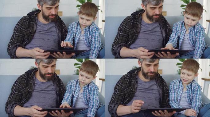慈爱的父亲和他可爱的小孩正在使用桌子触摸屏，坐在家里的沙发上聊天。现代科技和幸福家庭理念。