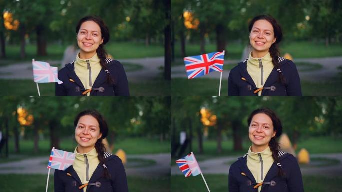 穿着便服的开朗年轻女士的慢动作肖像挥舞着英国的国旗，微笑着看着相机，背景是公园。
