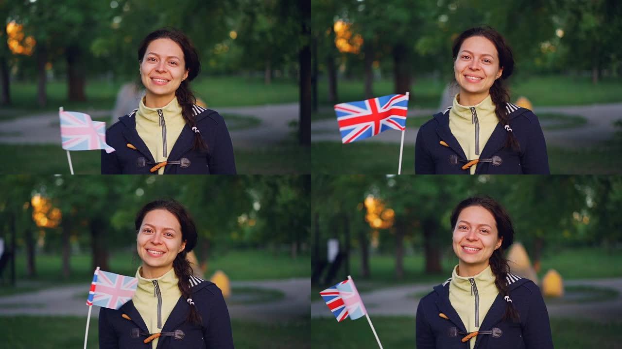 穿着便服的开朗年轻女士的慢动作肖像挥舞着英国的国旗，微笑着看着相机，背景是公园。