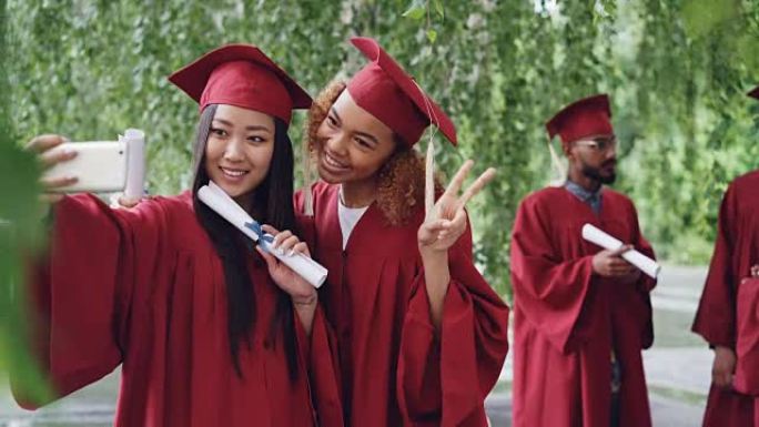 两个迷人的女孩同学在毕业典礼上拿着文凭自拍，年轻女性微笑着，摆姿势，做手势和滑稽的脸。