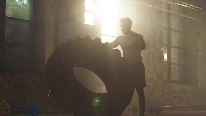 强壮肌肉的男人举起轮胎作为他健身/交叉健身训练的一部分。他满身是汗，在一个废弃工厂改造成的健身房锻炼