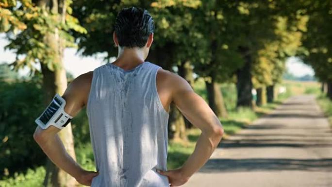 一个男人放松慢跑，在树木和大自然的街道上跑步，用耳机听音乐，在日落时跑得很慢。自然界中运动和健身放松
