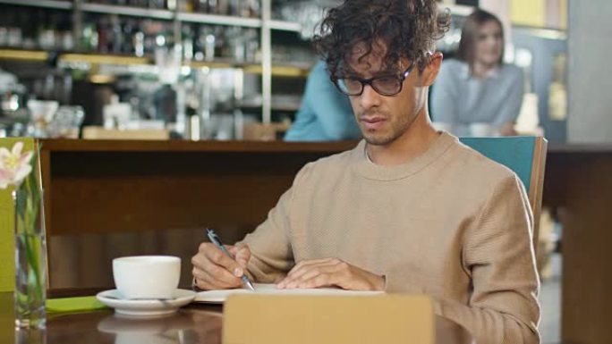 西班牙裔年轻人在舒适的咖啡店使用平板电脑并在笔记本上做笔记。