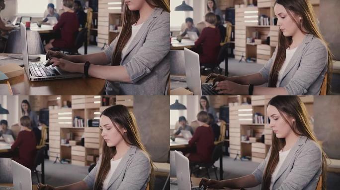 集中的欧洲年轻女孩在笔记本电脑上打字。在时尚的办公空间中努力工作，在背景中团队合作4K