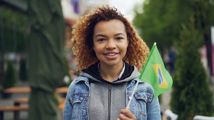 快乐的非洲裔美国女孩看着相机，举着巴西国旗站在现代城的尼斯公园里的慢动作肖像。旅游和人的概念。