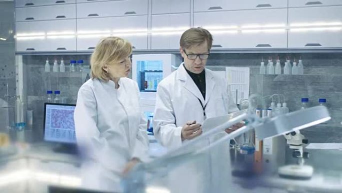 女性和男性首席科学家走过现代实验室，讨论未来的可能性。年轻的女助理在她的办公桌前工作。