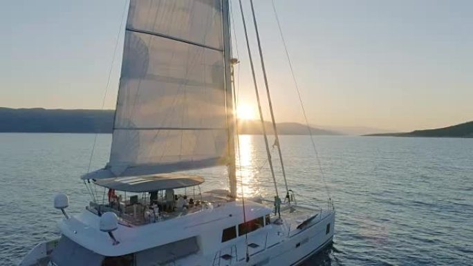 一艘帆船双体船的空中拍摄，帆船在平静的海面上航行，太阳升起在山后。