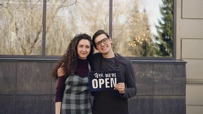 幸福的已婚夫妇商业伙伴的肖像开咖啡馆，在大楼外的窗前拿着 “我们是开放的” 板。成功的创业理念。