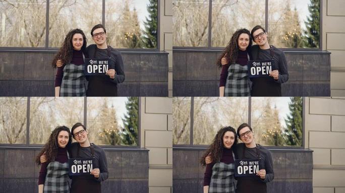 幸福的已婚夫妇商业伙伴的肖像开咖啡馆，在大楼外的窗前拿着 “我们是开放的” 板。成功的创业理念。